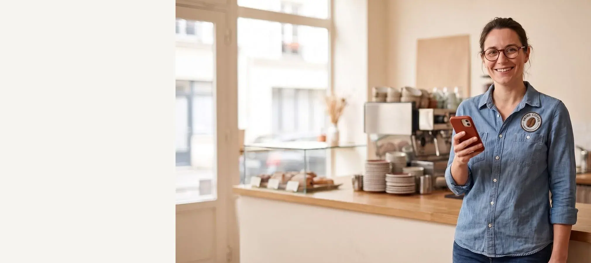 Smiling barista in a chambray shirt with a red-cased phone in hand, neighborhood cafe behind her