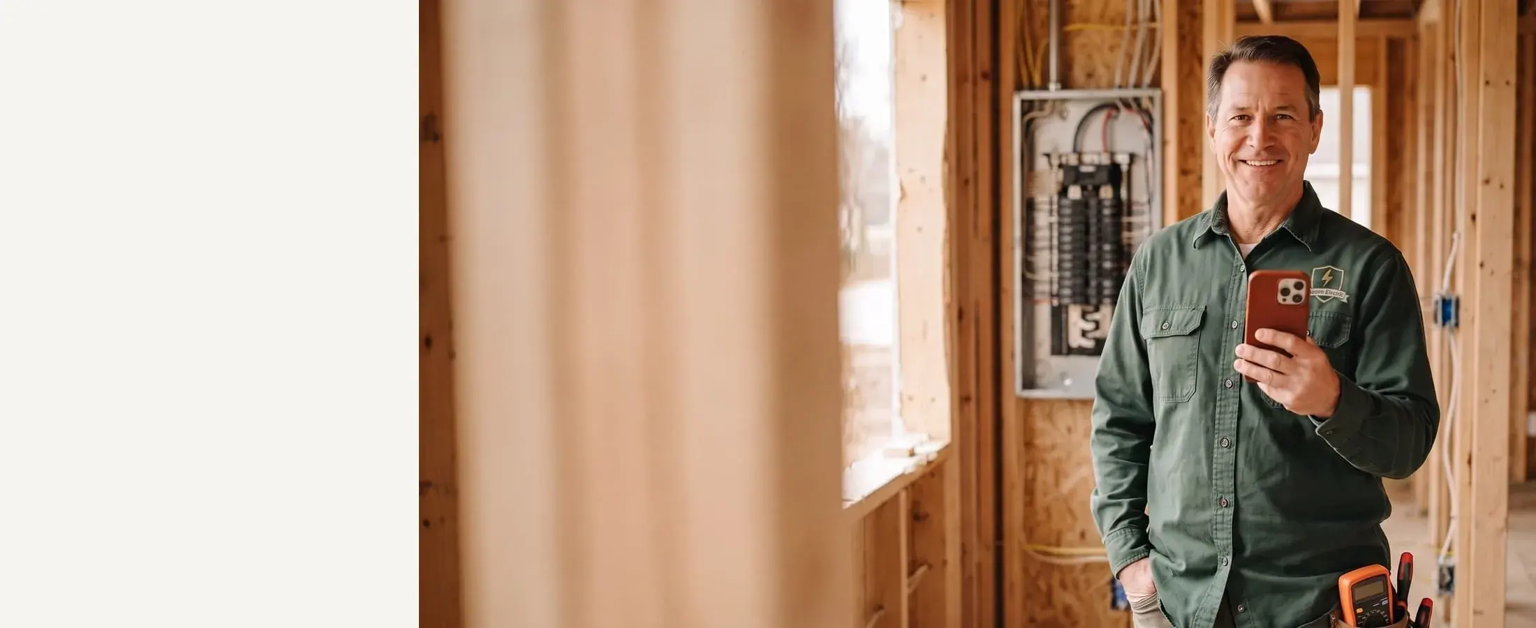 Smiling Beacon Electric electrician in a hunter-green shirt with a red-cased phone, open breaker panel behind him