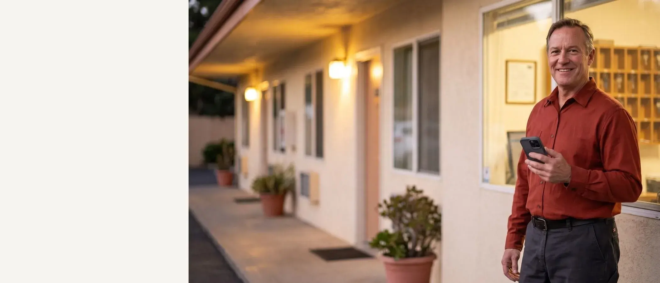Smiling motel manager in a rust-red shirt with a phone in hand, motel walkway at dusk behind him