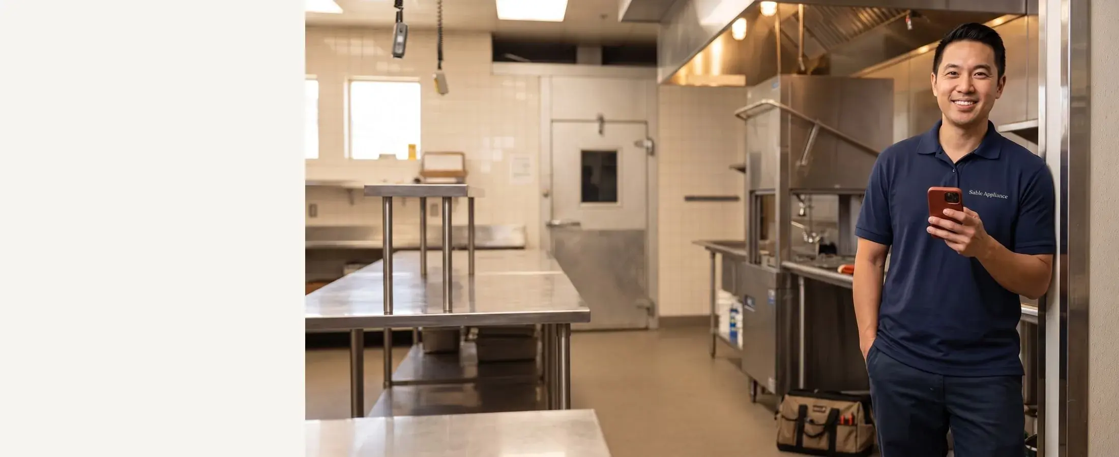 Smiling Sable Appliance repair technician in a navy polo with a red-cased phone, an open commercial dishwasher in the restaurant kitchen behind him