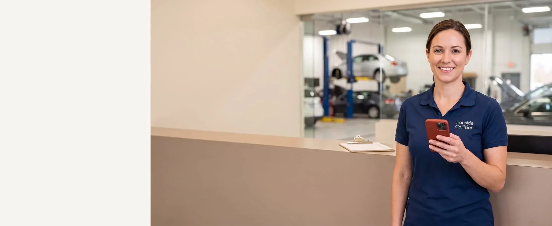 Smiling auto body service advisor in a navy polo with a red-cased phone in hand, glass-walled repair shop behind her
