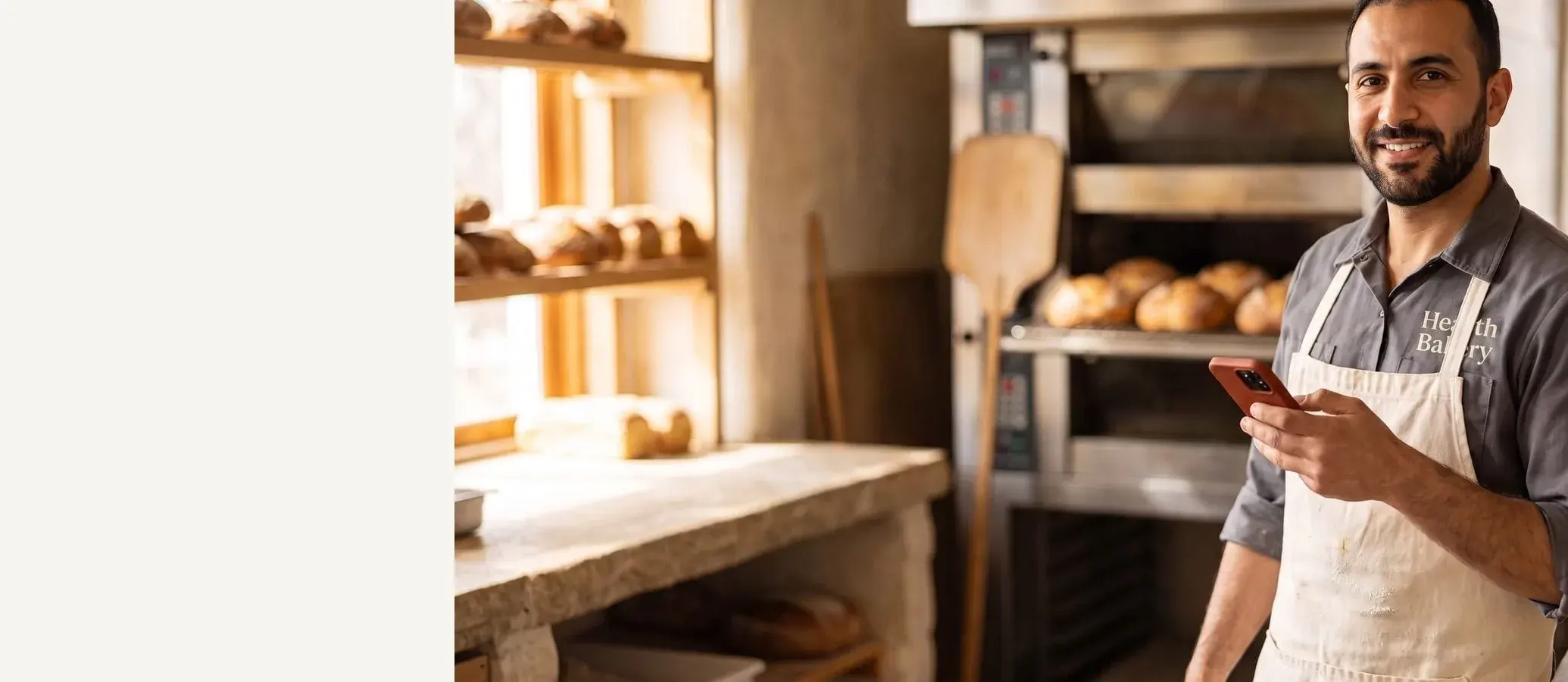 Smiling baker in a bib apron with a red-cased phone in hand, warm bakery kitchen behind him