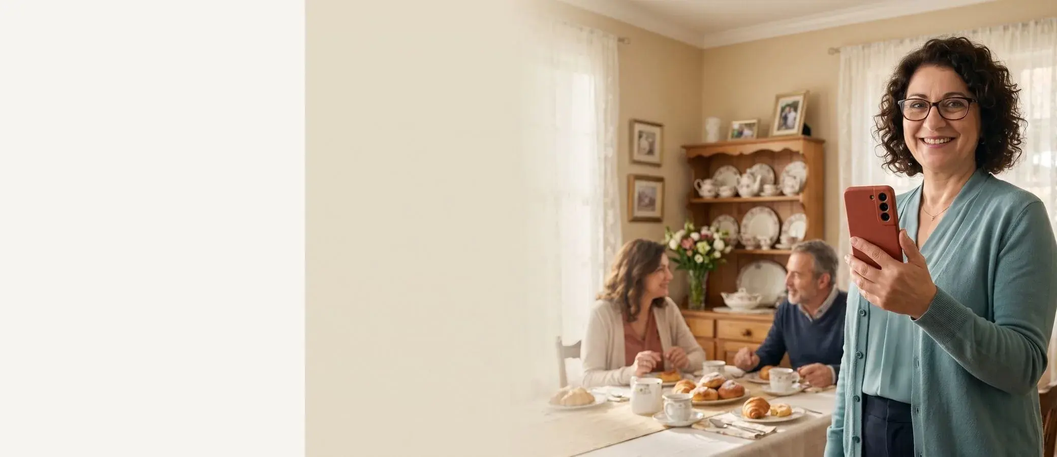 Smiling bed and breakfast innkeeper with curly hair and glasses, red-cased phone in hand, breakfast dining room with guests behind her