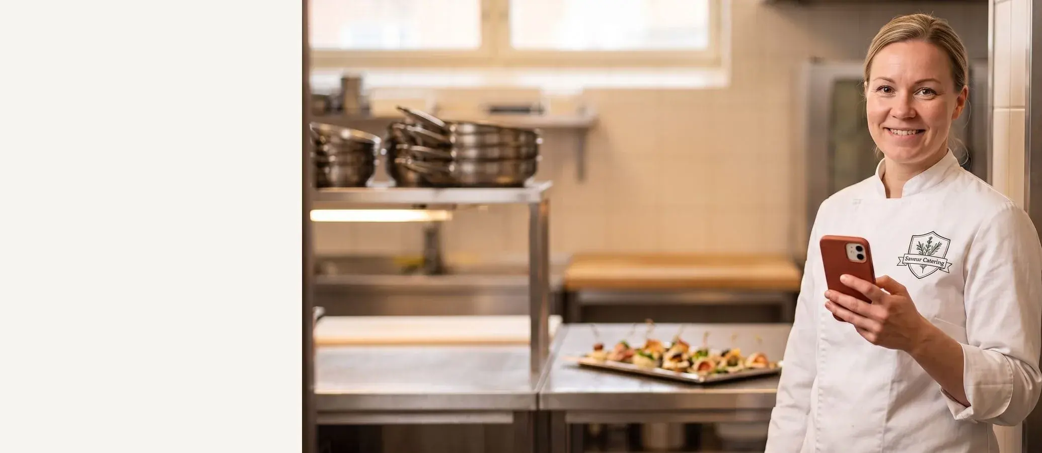 Smiling catering chef in a white chef coat with a red-cased phone in hand, clean catering kitchen behind her