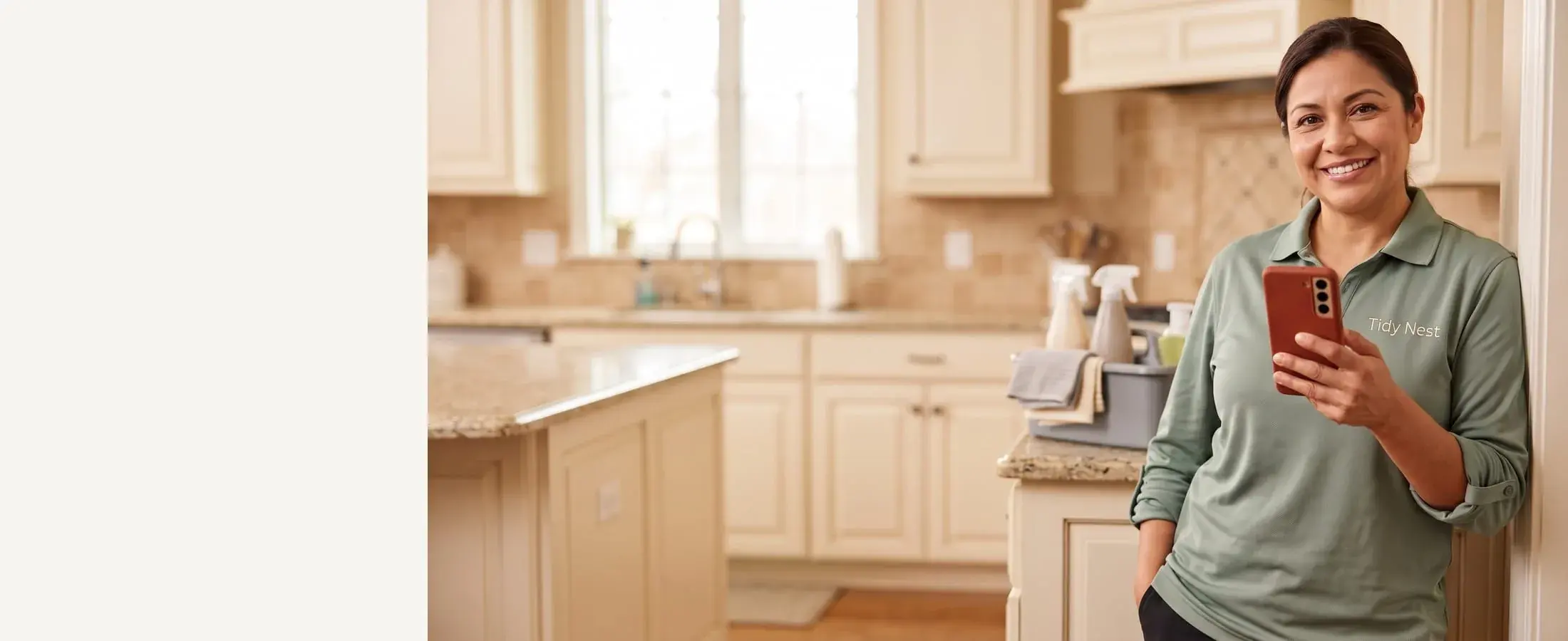 Smiling Tidy Nest house cleaner in a sage polo with a red-cased phone, tidy kitchen behind her