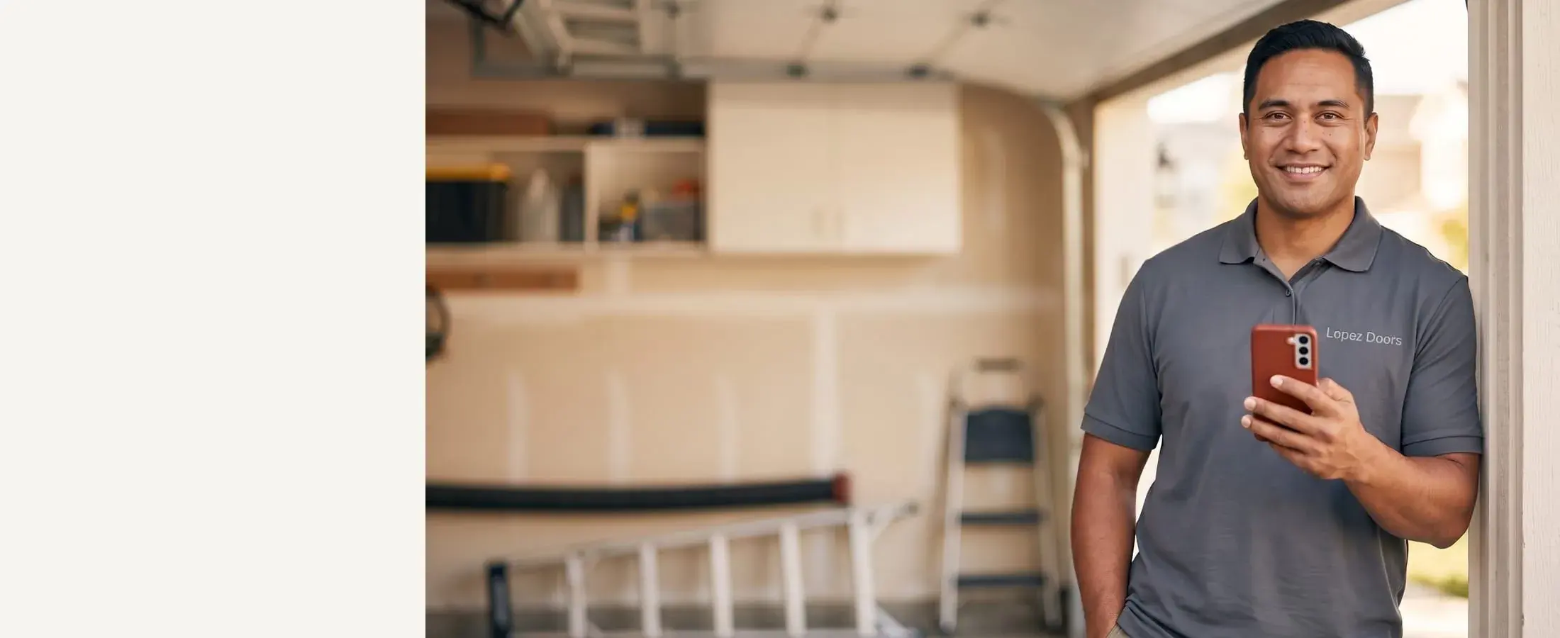 Smiling Lopez Doors technician in a deep stone-gray polo with a red-cased phone, open garage door track behind him