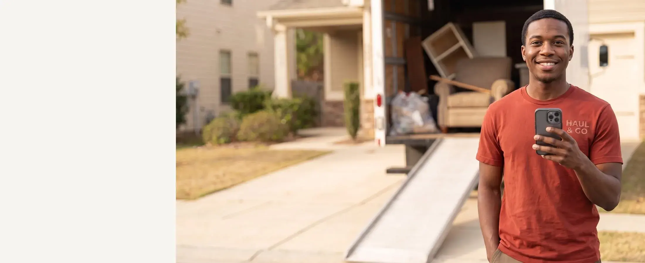 Smiling Haul & Go junk removal crew member in a red tee with a phone in hand, loaded box truck behind him