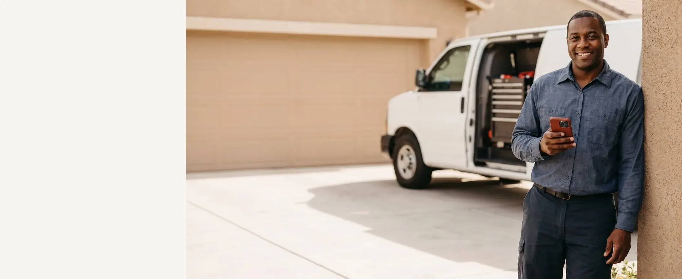 Smiling mobile mechanic in a chambray work shirt with a red-cased phone in hand, service van at the curb behind him