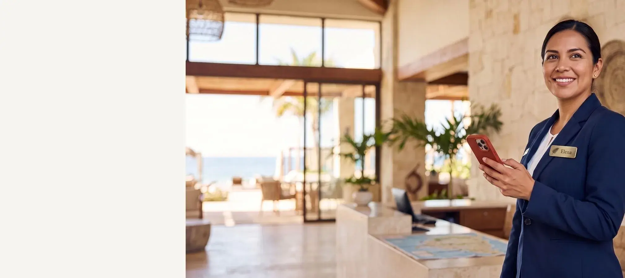Smiling resort concierge in a navy blazer with a gold "Elena" name badge and a red-cased phone in hand, beach resort lobby behind her