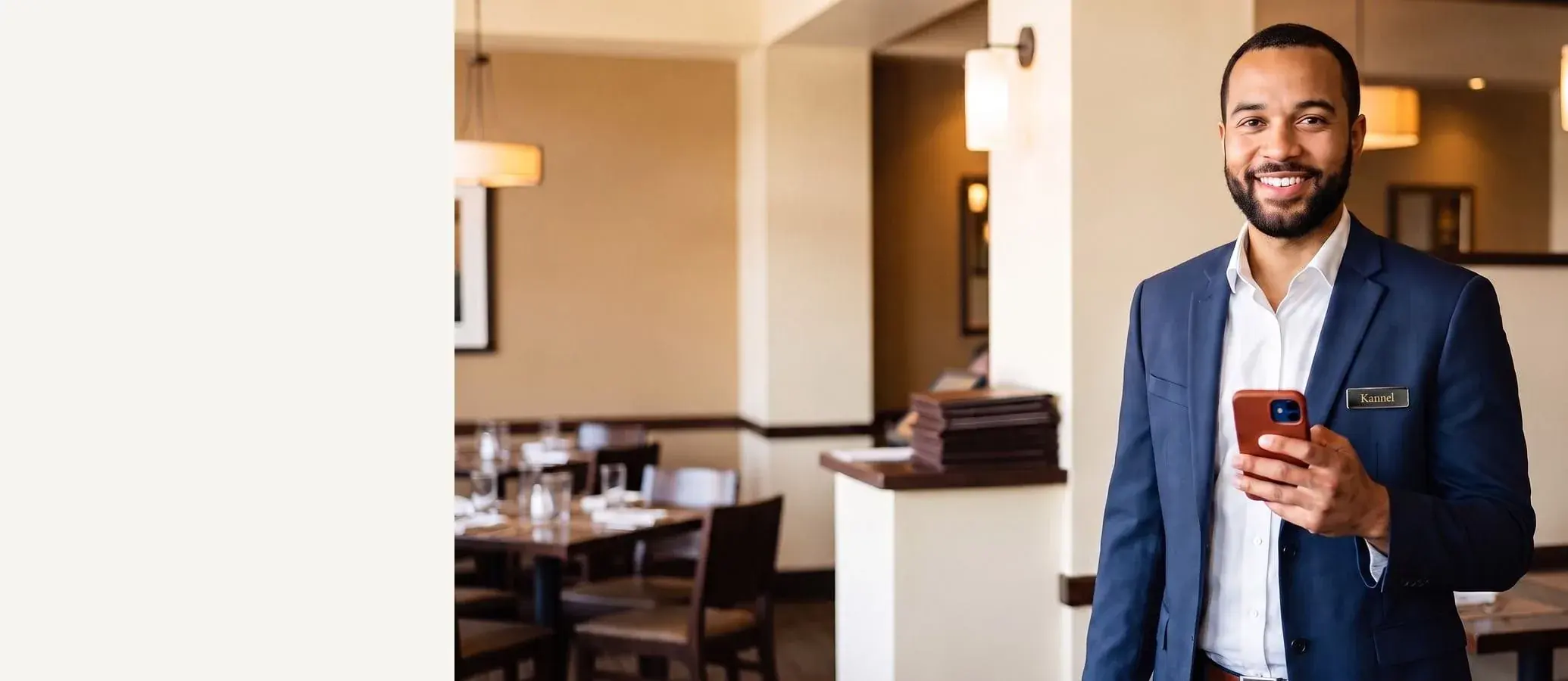 Smiling restaurant manager in a navy blazer over a crisp white shirt with a Kannel name tag and a red-cased phone in hand, elegant dining room behind him