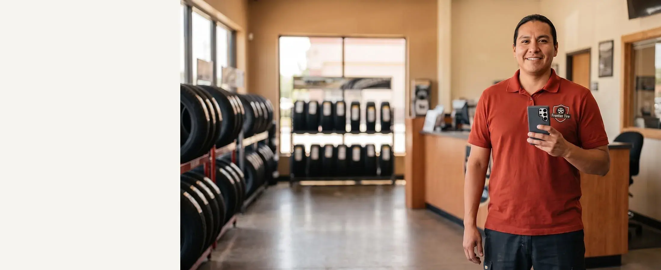Smiling tire shop advisor in a rust-red polo with a phone in hand, stocked tire racks behind him